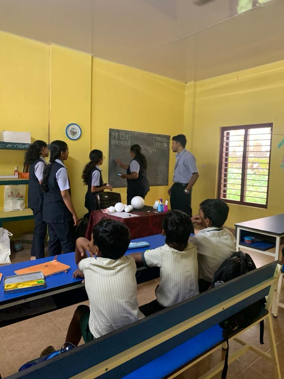 Student volunteers writing on the blackboard at the front of a yellow classroom while children watch from wooden benches.