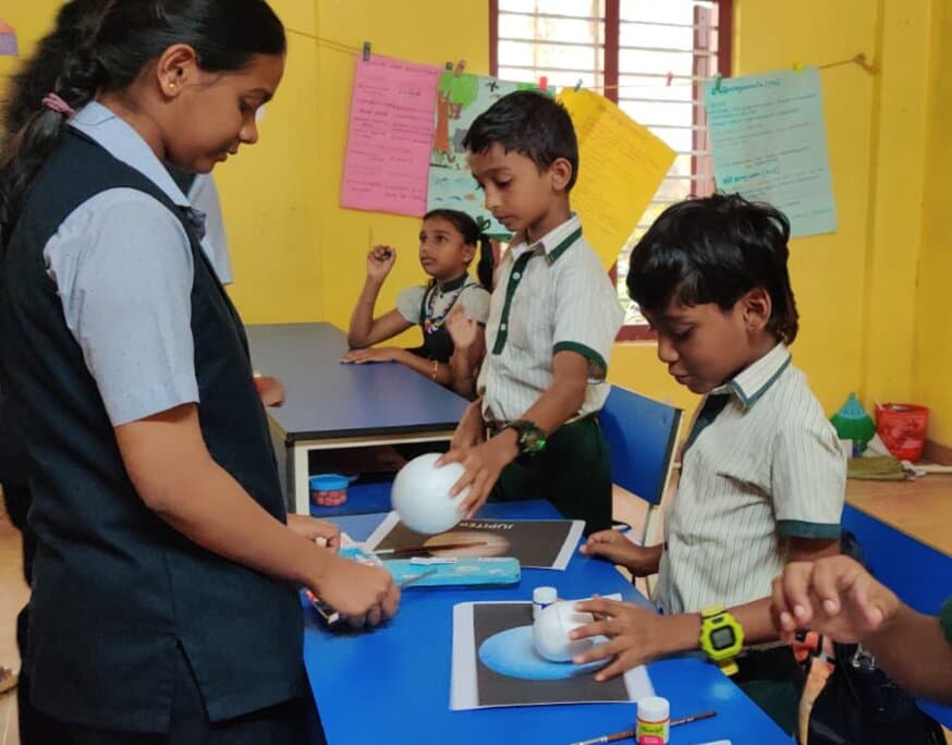Children and a volunteer assembling white polystyrene spheres into planet models, with glue, brushes and a Jupiter reference card on the desk.