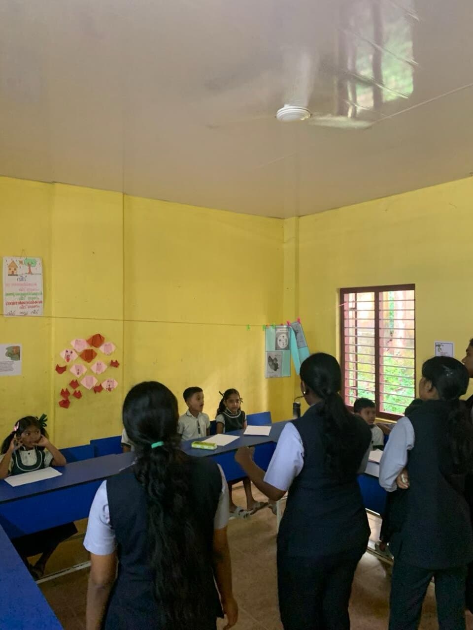 Volunteers standing among the desks of a bright yellow classroom, working with young children seated at blue benches.