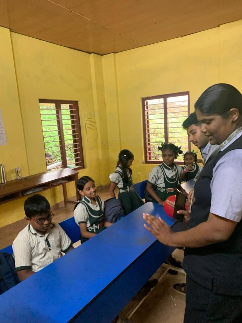 A student volunteer holding up a red ball to demonstrate a concept while children gather around the classroom bench.