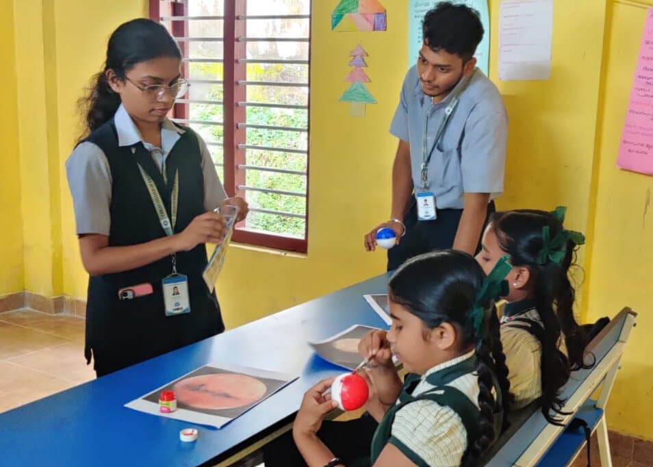 A student volunteer holding a reference card while a young girl paints a polystyrene sphere red to make a model of Mars.
