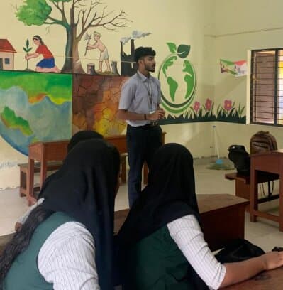 A student volunteer presenting to seated listeners in front of a painted wall mural of the earth and environmental scenes.