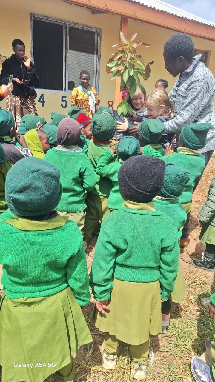 A crowd of children in green uniforms outside the school, gathered around adults handing over a young potted tree seedling.