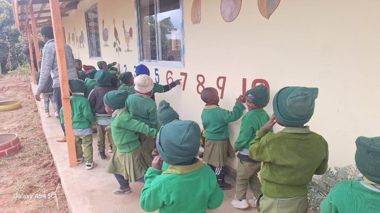 Children lined up along a veranda reaching up to touch large painted numerals one to ten on the school wall.