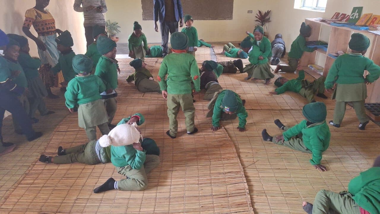 Children spread out across floor mats mid-game, stretching, crawling and lying down, with a shelf of learning materials against the wall.