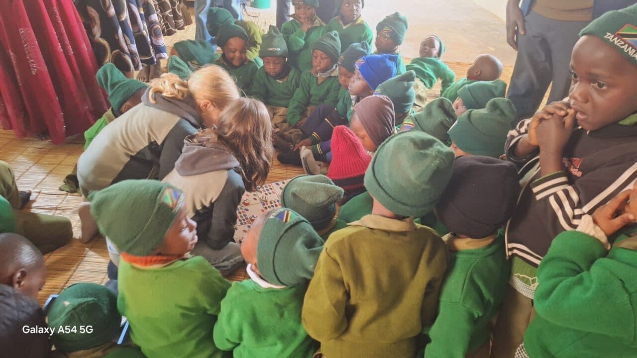 A large group of children in green school jumpers and knitted hats crowded around adults working with number cards on a woven floor mat.