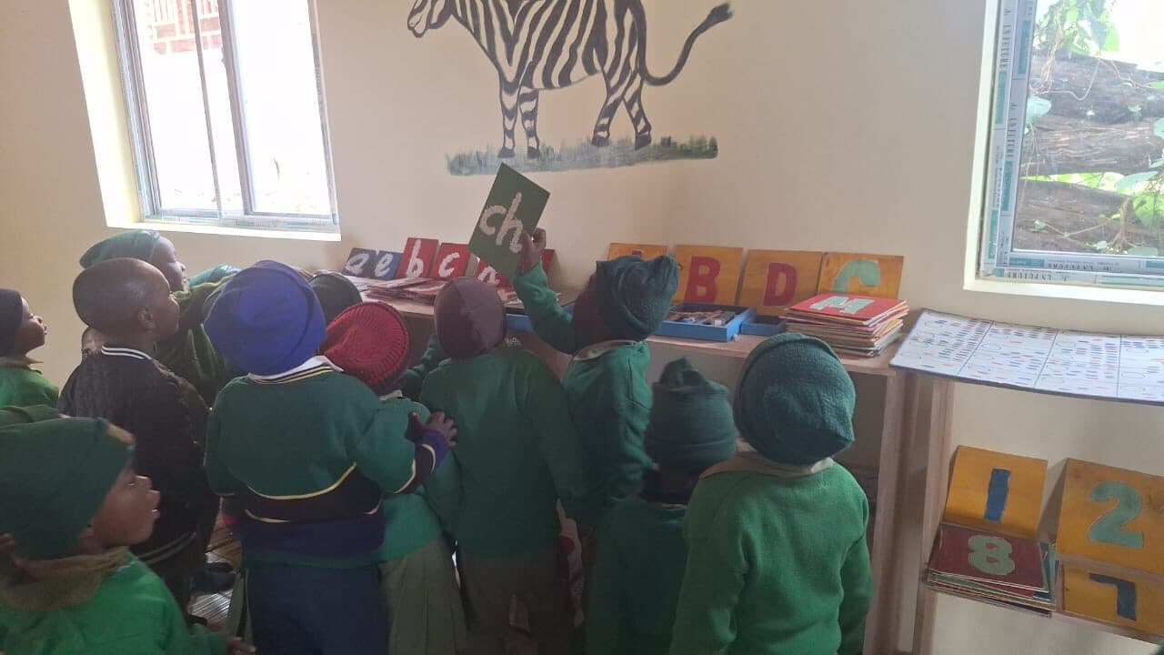 Children standing at a low wooden shelf of painted letter cards, one child holding up a green card reading ch.