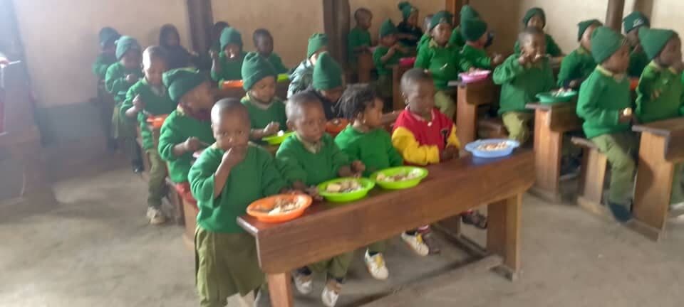 Children seated at wooden benches eating from coloured plastic bowls in a classroom.