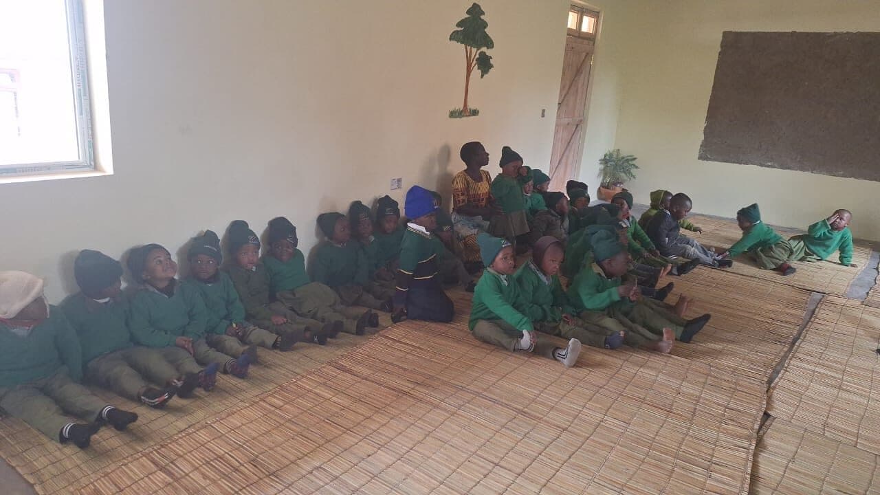 Children sitting in rows along the walls of a bare classroom on woven mats, listening, with a teacher seated among them.