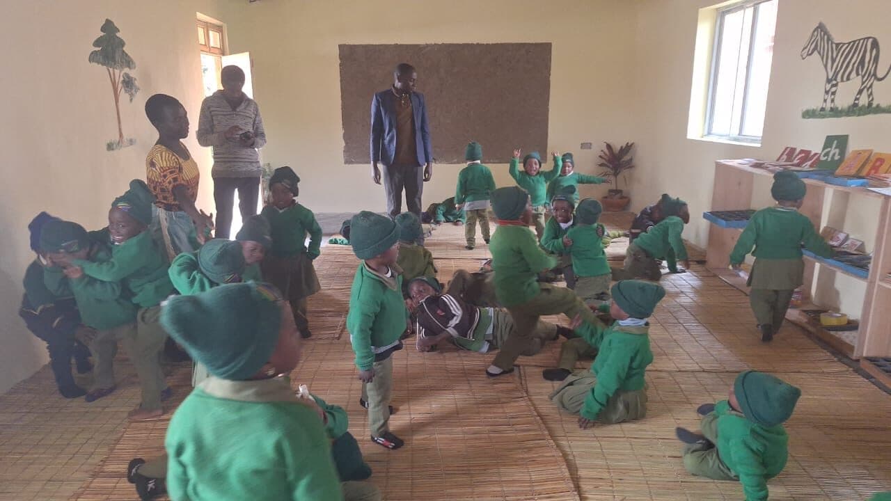 Children moving freely across the woven floor mats of a classroom, some running, some rolling, with adults watching from the side.