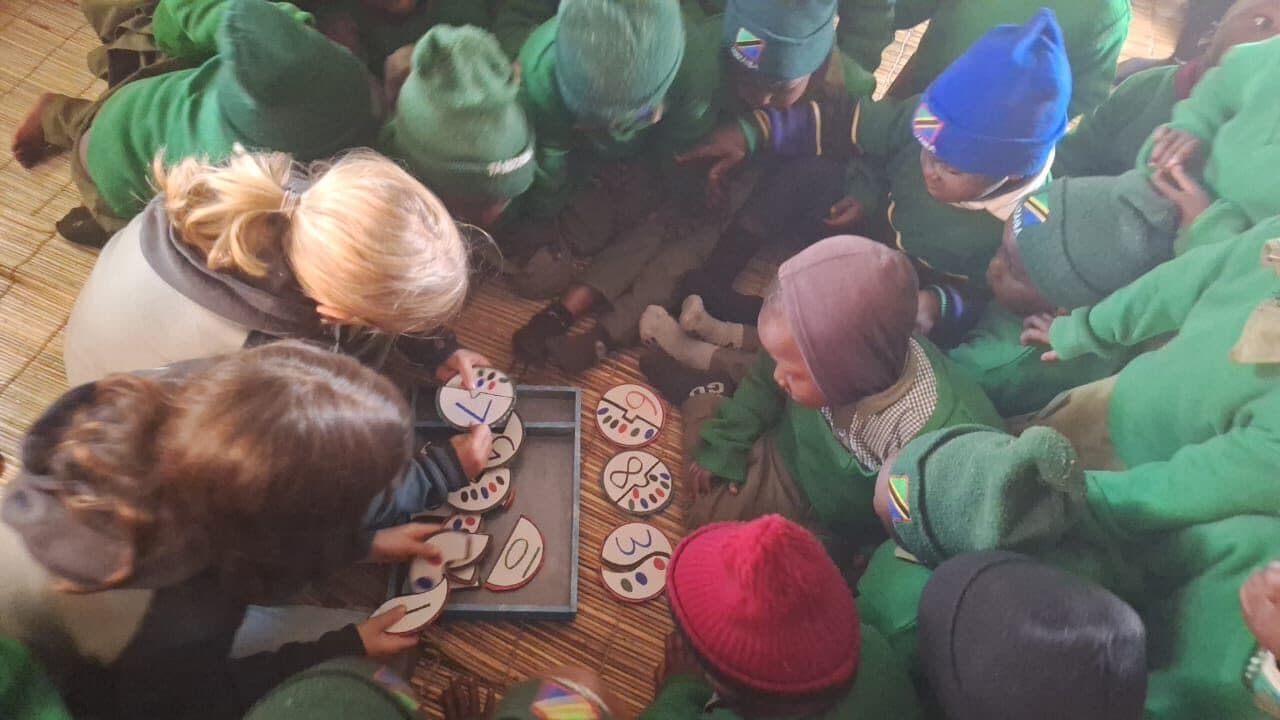 Adults and children kneeling around numbered discs and a shallow tray on a woven mat, arranging them into order.