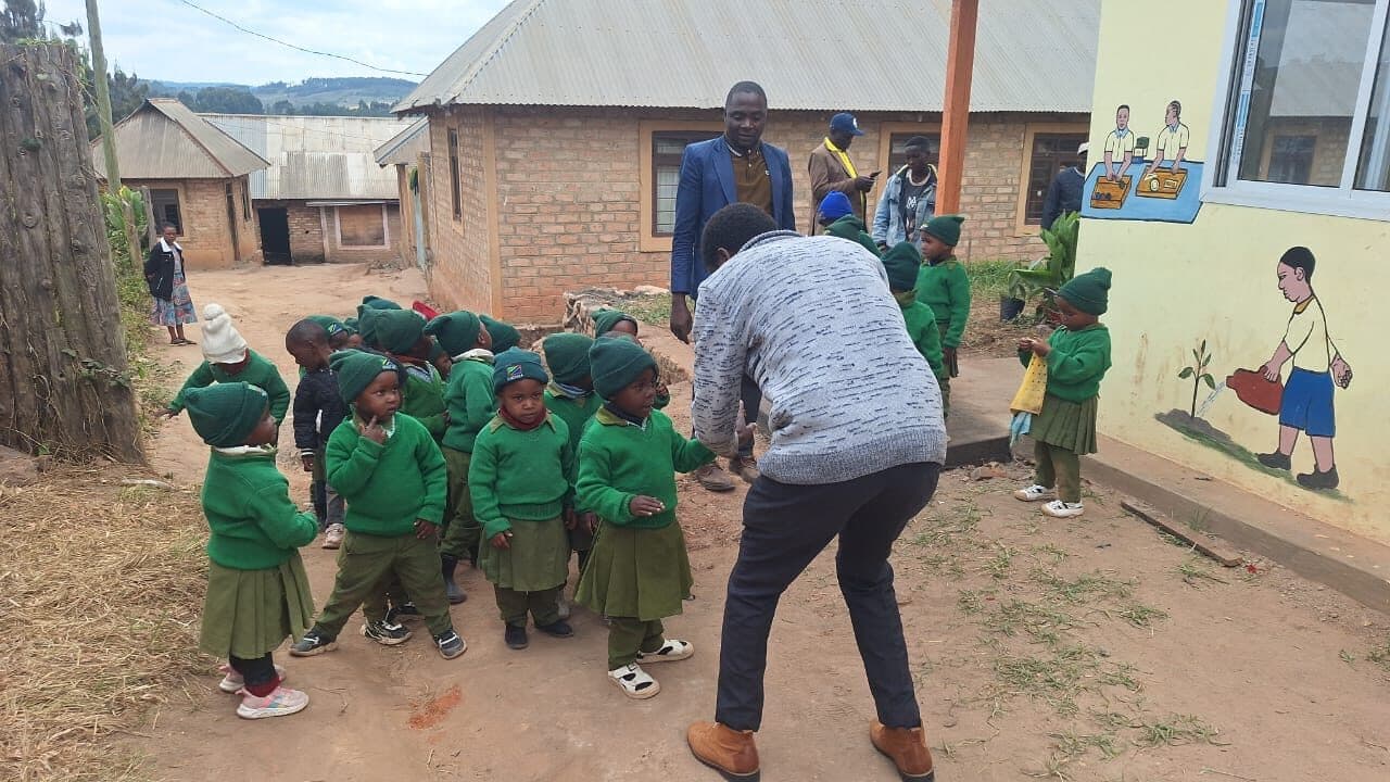 Children in green uniforms gathered outside brick school buildings on a dirt path, with a teacher crouching to speak with them and hills in the distance.