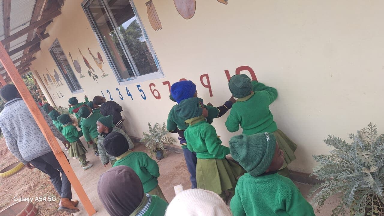 Young children stretching up on tiptoe to reach painted numbers high on the veranda wall, with painted birds and animals above the windows.