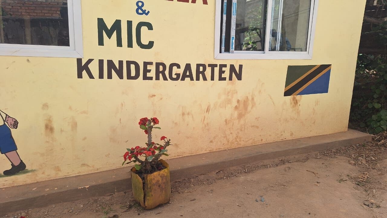 The painted exterior wall of a kindergarten building, lettered MIC KINDERGARTEN beside a painted Tanzanian flag, with a potted flowering plant below.