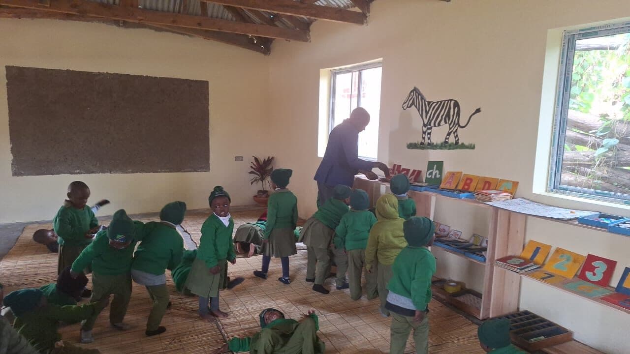 A classroom with woven floor mats, a mud-plastered blackboard, a painted zebra on the wall and a low wooden shelf holding numbered and lettered cards.