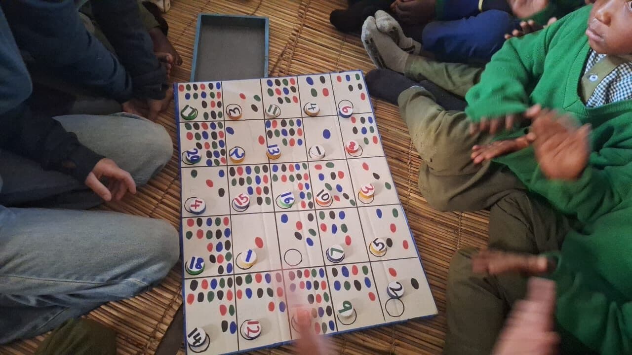 A hand-made counting board of numbered discs and coloured dots laid on a woven mat, with children sitting around it clapping.