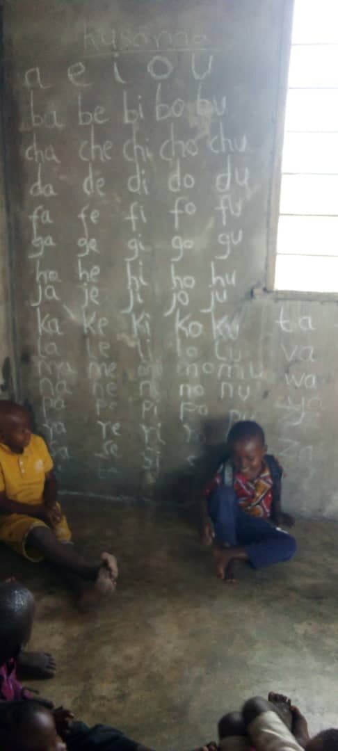 A Swahili syllable chart headed Kusoma, chalked in rows onto a bare concrete wall, with two small children sitting on the floor in front of it.