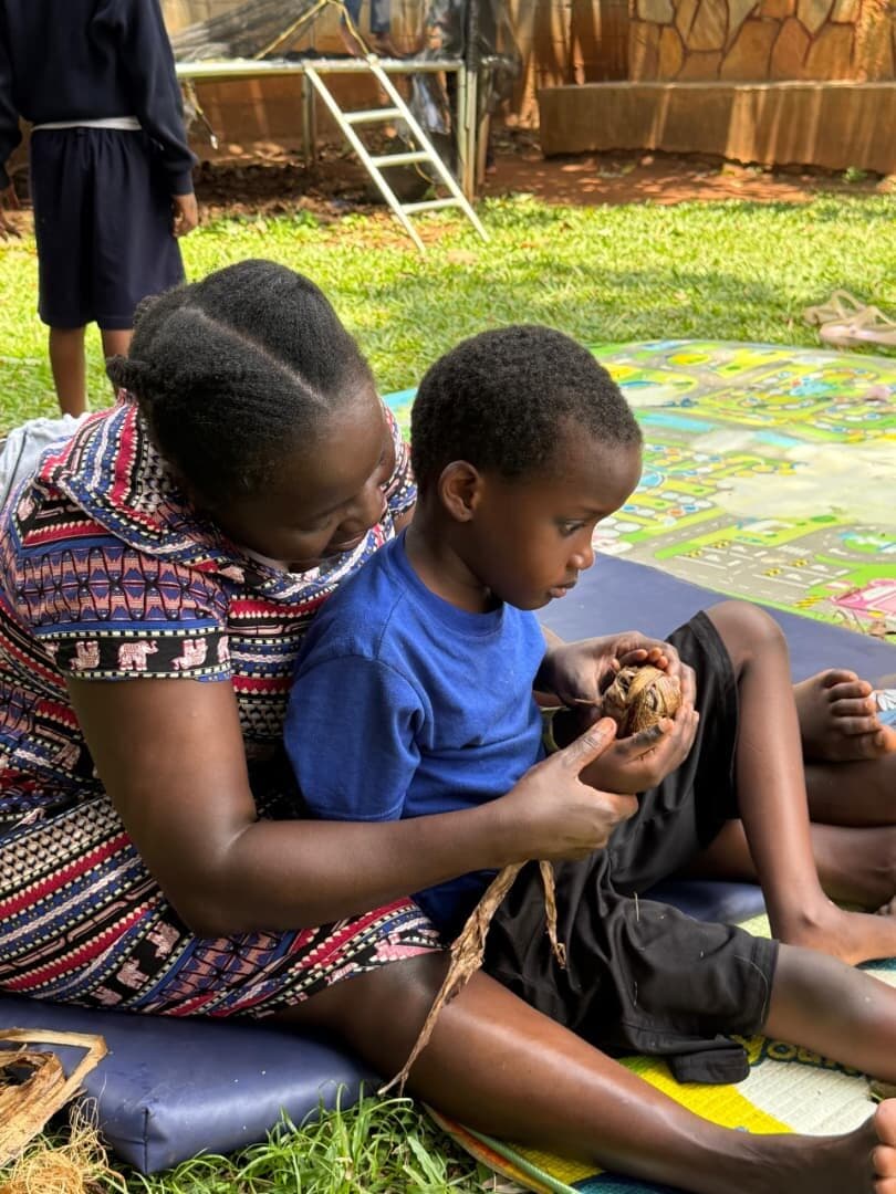 A teacher holding a young learner on her lap, the two of them shaping a small banana fibre doll together.