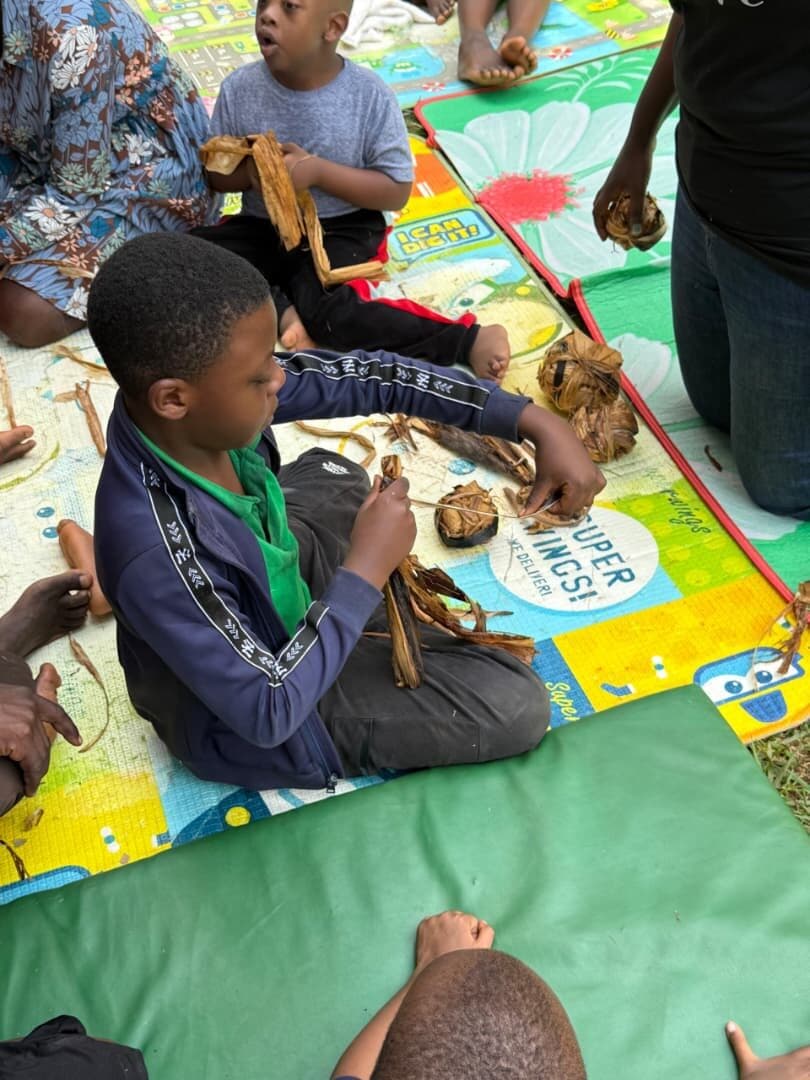 A learner sitting cross-legged on a play mat, concentrating on binding strands of banana fibre together.