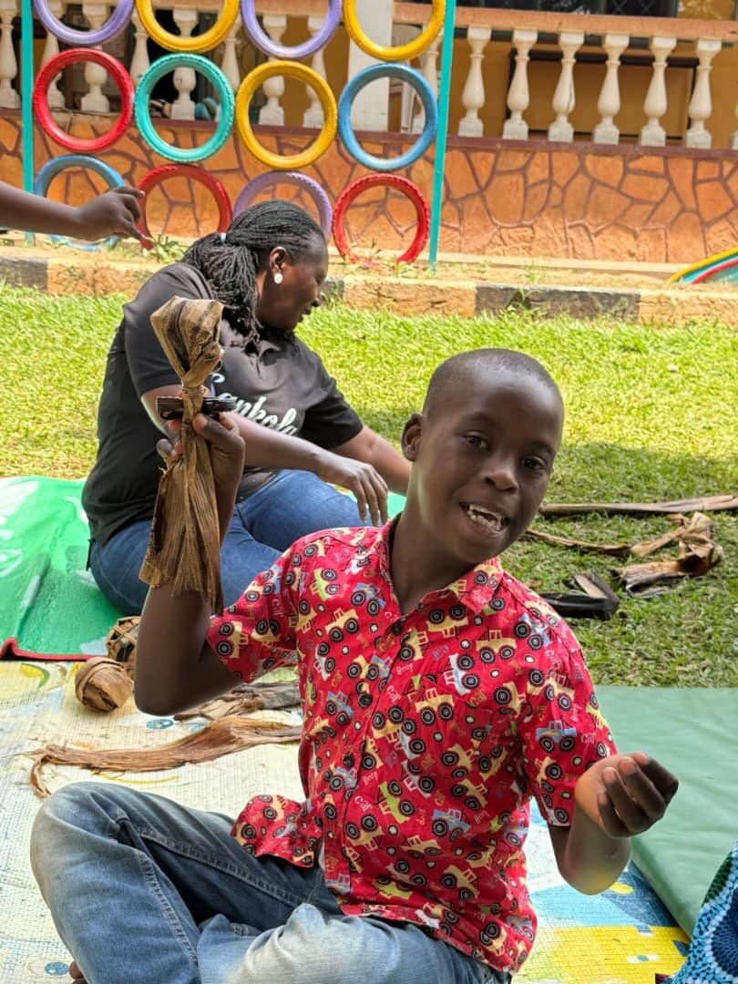 A smiling learner holding up a banana fibre doll he made, sitting on a mat in the compound.