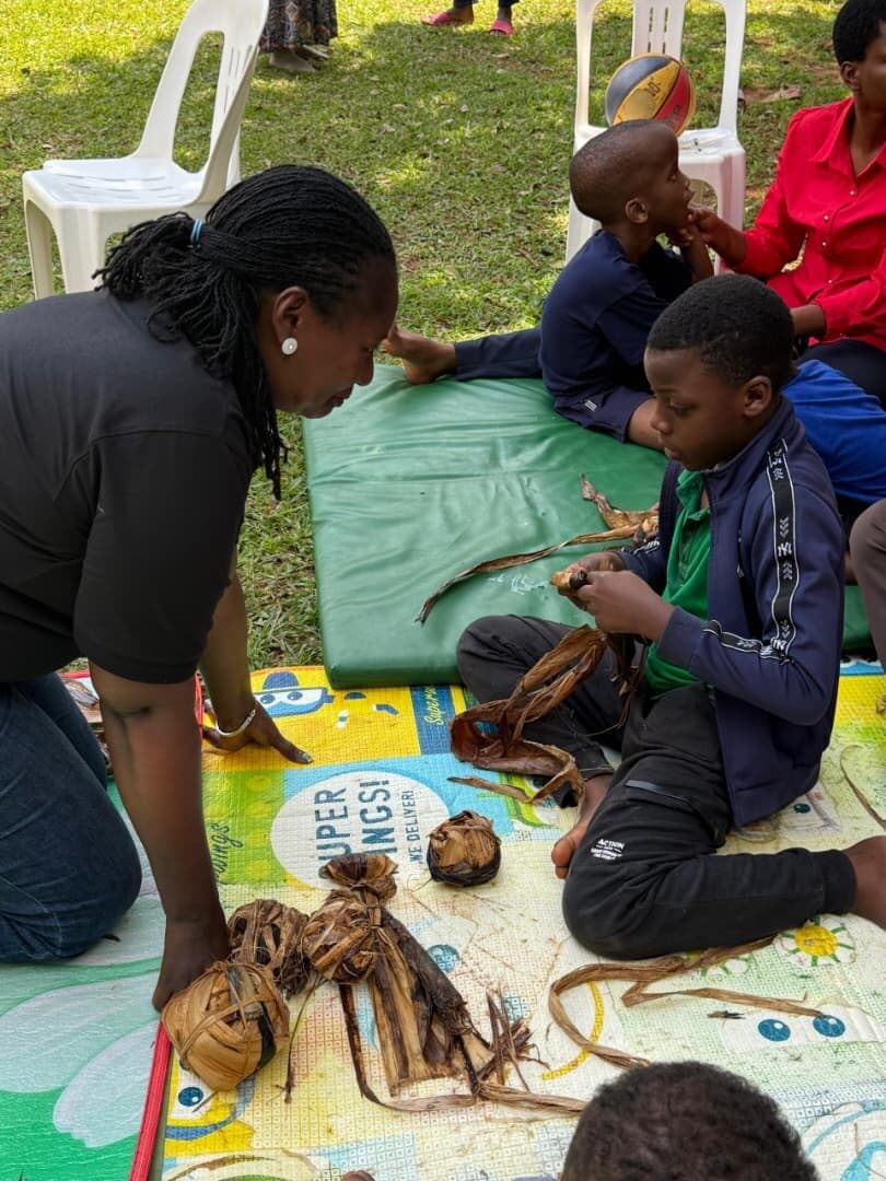 A teacher and a learner working side by side on the mat, surrounded by finished banana fibre creations.