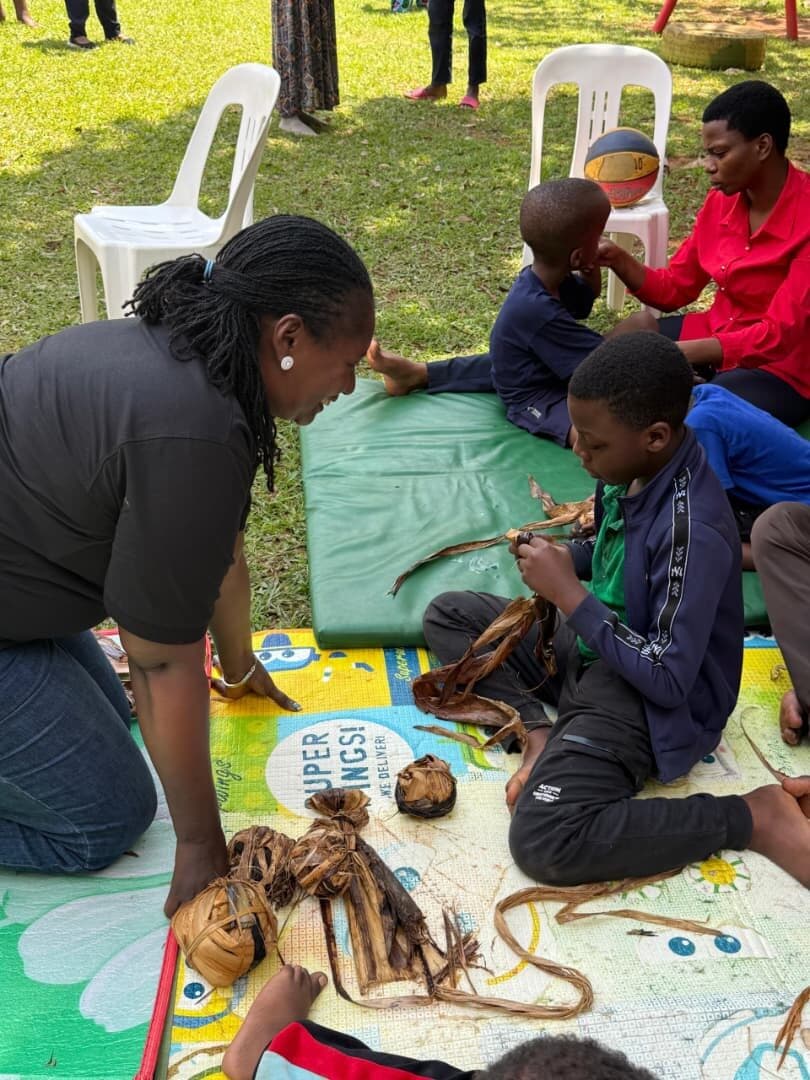 A teacher leaning in beside a learner on the mat, guiding their hands as they work with banana fibre.